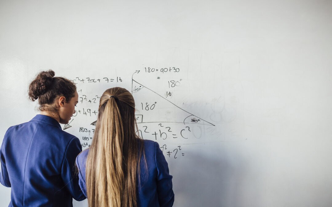 Two teenage school girls standing in front of a large whiteboard side by side solving a mathematics equation on the board using computational thinking. Back view