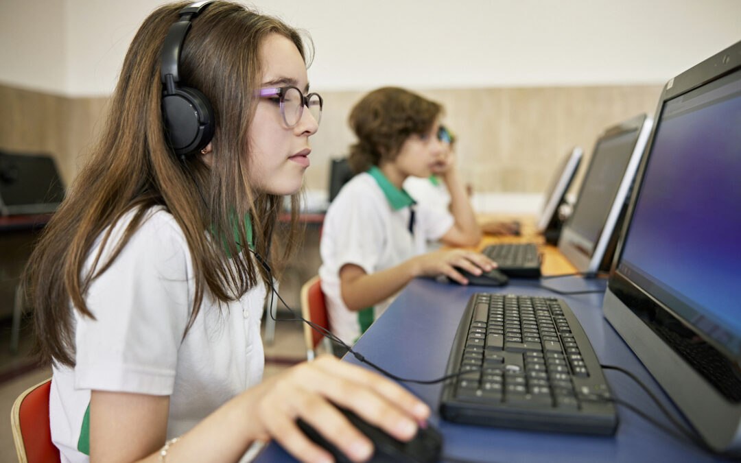 Focus on foreground 11 year old girl with long hair, wearing eyeglasses, headphones, and using mouse as she completes a digital literacy assessment on a computer