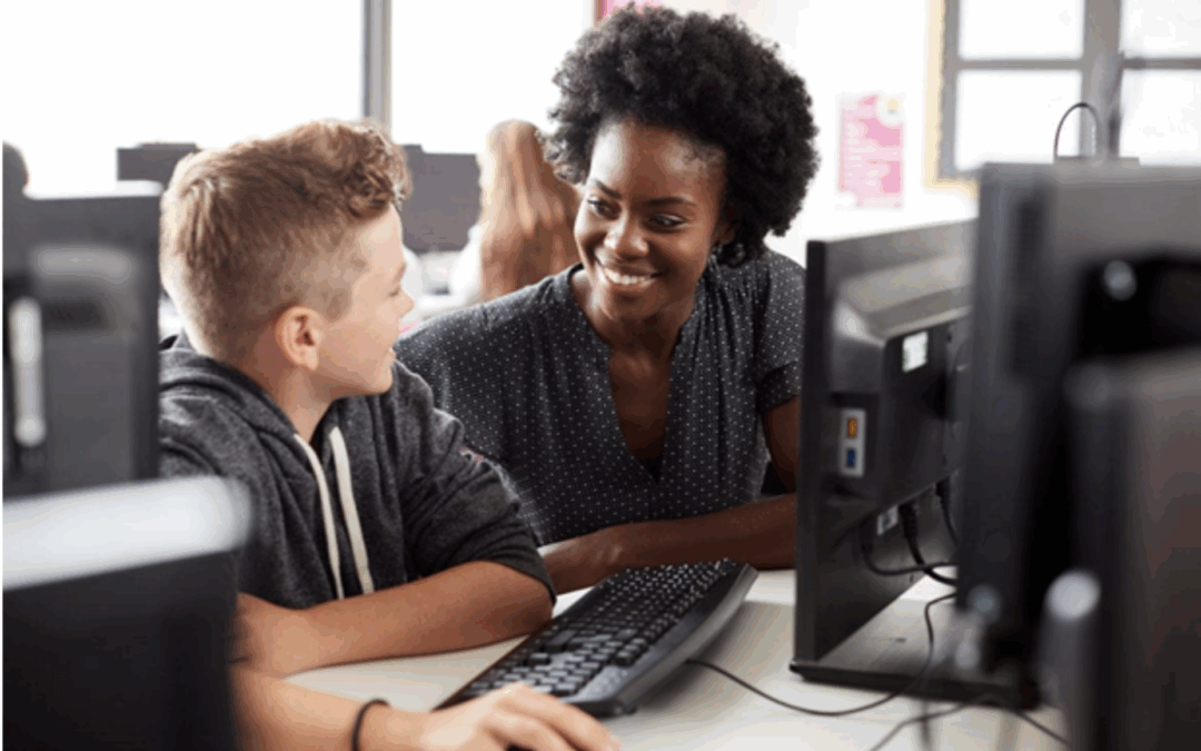 Smiling teacher assisting a student at a computer in a classroom setting